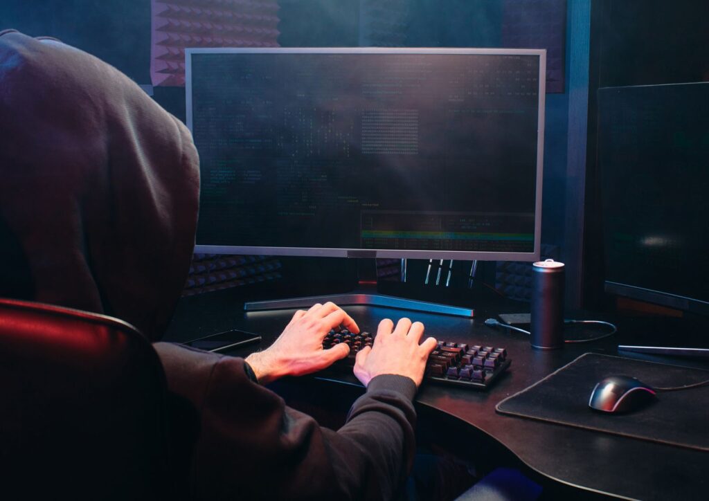 A hooded figure seen from behind facing a monitor with code, illustrating state-sponsored cyber attack