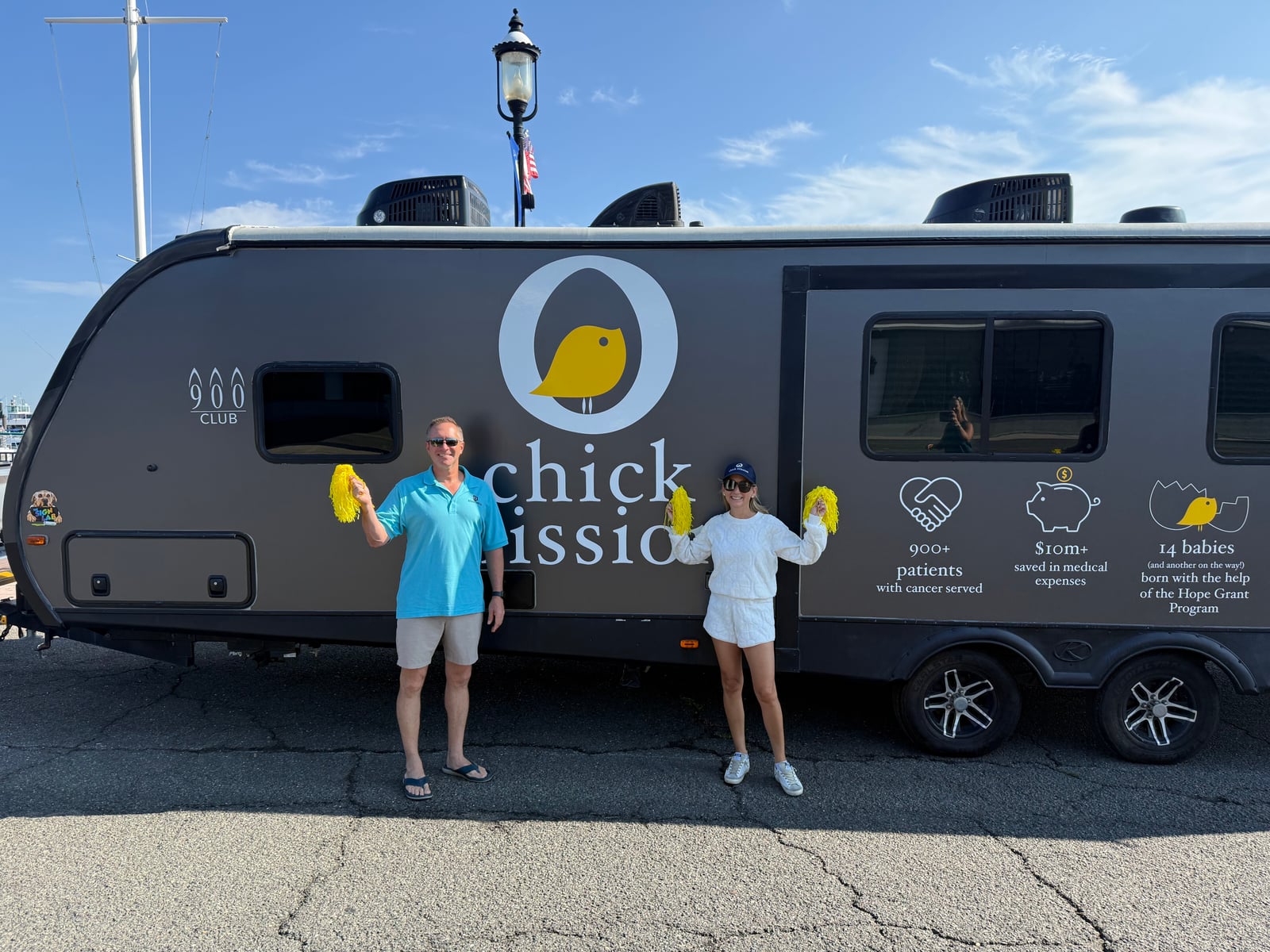 Glenn Kupsch of Adaptive IT stands with a Chick Mission supporter in front of the chick camper's logo and Hope Grant statistics, both holding yellow pom-poms.