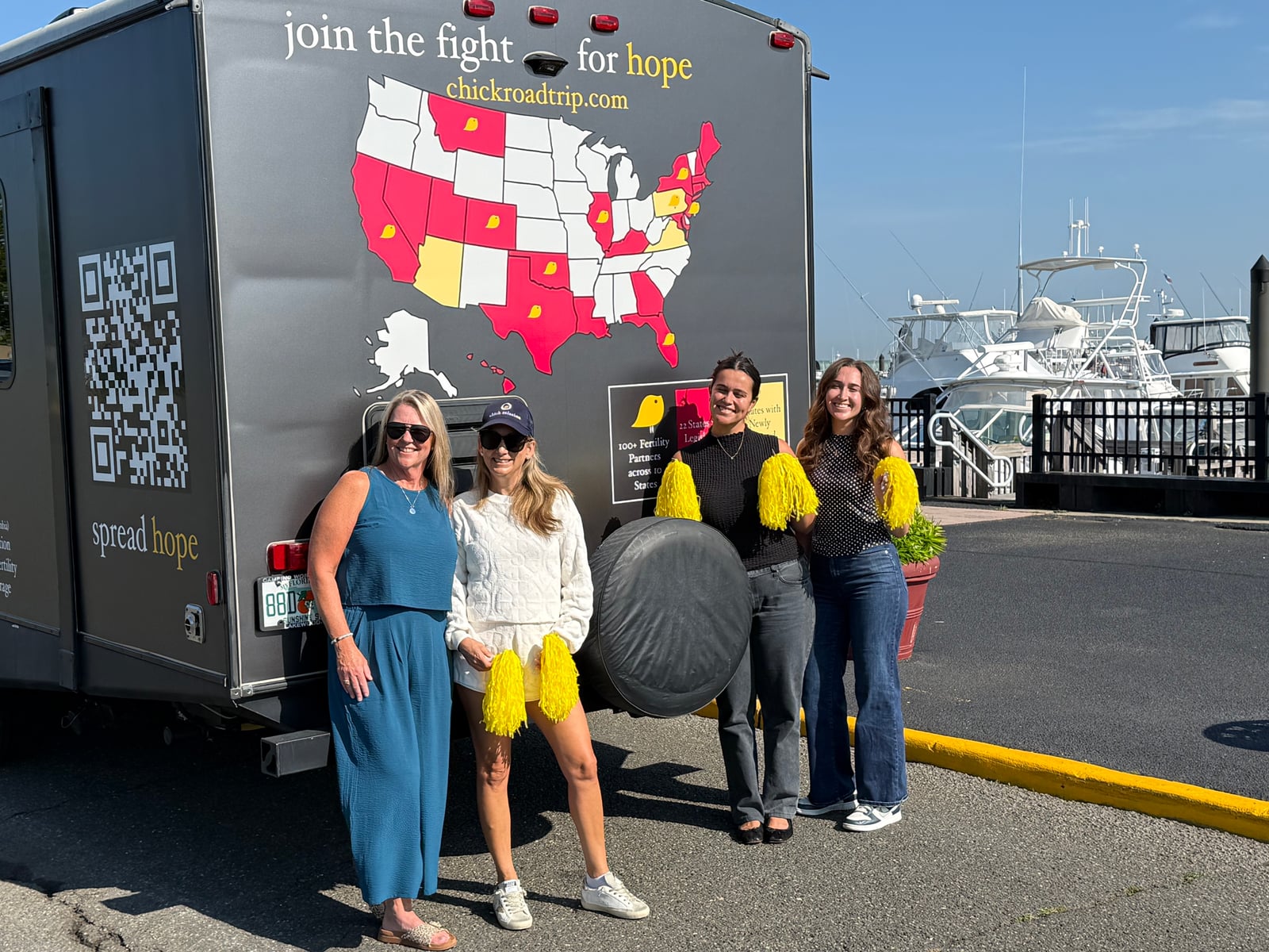 Rear of The Chick Mission road-trip camper showing a red-and-yellow US map, chickroadtrip.com, and four supporters holding yellow pom-poms in front of the Atlantic Highlands marina.